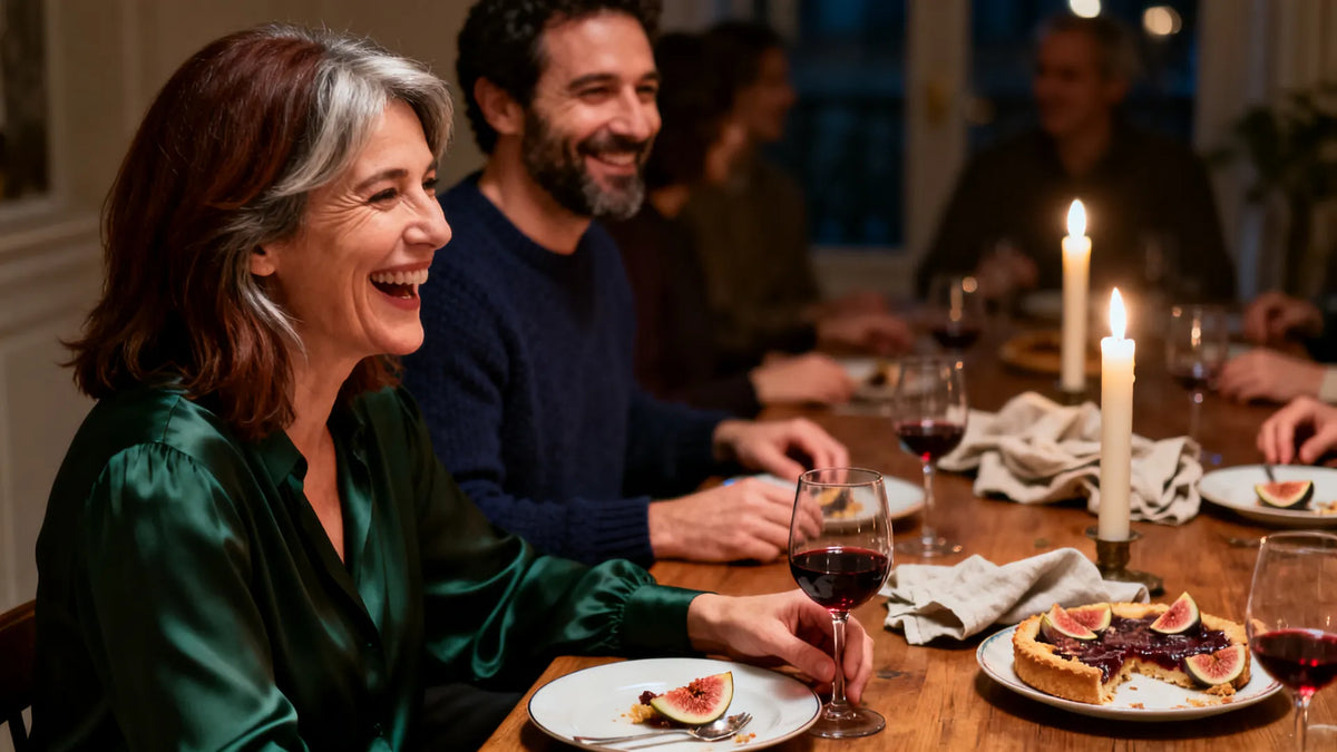 Fin de dîner aux chandelles autour d'une table en bois : une femme d'une cinquantaine d'années à la mèche grise rit, un verre à la main, à côté d'un homme barbu en pull marine, devant une tarte aux figues entamée.